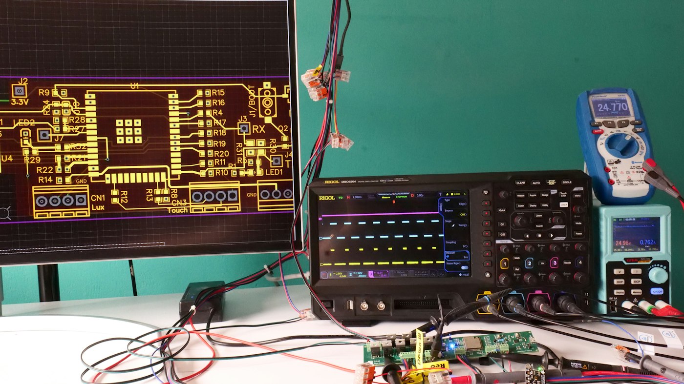 An engineering bench with a PCB layout on a monitor, an oscilloscope and a prototype board wired up for measurement